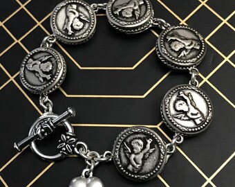 May include: Silver-toned bracelet featuring round charms with embossed cherub and mythological figure designs. The bracelet has a toggle clasp and a heart charm. The bracelet is displayed on a black surface with a gold grid pattern.