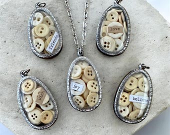 May include: A silver-toned necklace with a pendant filled with small, off-white buttons. Four additional pendants with buttons are arranged around the necklace. Each pendant has a small label with words like "friends", "love", "wish", "joy", and "believe".
