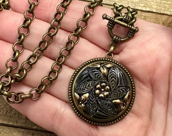 May include: A gold chain necklace with a large, round pendant. The pendant is black with a gold floral design.