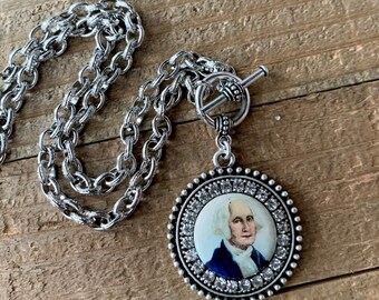 May include: A silver chain necklace with a silver toggle clasp. The pendant is a round silver frame with a white background and a portrait of George Washington. The frame is decorated with clear rhinestones.