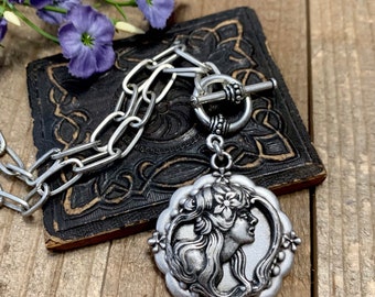 May include: A silver necklace with a toggle clasp and a pendant featuring a detailed profile of a woman with flowers in her hair.