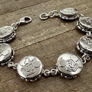 May include: A silver bracelet featuring circular charms with embossed images of Mickey and Minnie Mouse. The bracelet has a lobster clasp closure and is displayed on a wooden surface. The charms are approximately 0.75 inches in diameter.