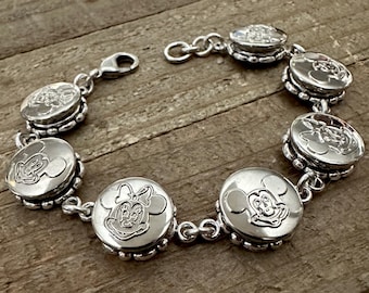 May include: A silver bracelet featuring circular charms with embossed images of Mickey and Minnie Mouse. The bracelet has a lobster clasp closure and is displayed on a wooden surface. The charms are approximately 0.75 inches in diameter.