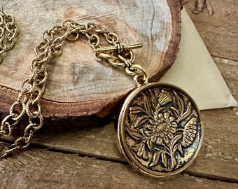 May include: A gold chain necklace with a large, round pendant featuring an embossed floral design. The pendant has a detailed, intricate design of flowers and leaves.