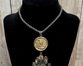 May include: A silver chain necklace with a gold pendant featuring a floral design and a dangling frog with red eyes. The frog is covered in black rhinestones.