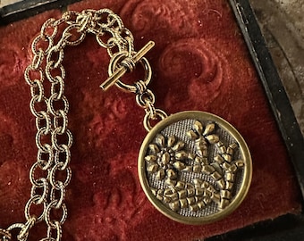 May include: Gold-toned necklace with a round pendant featuring a floral design. The pendant is attached to a double-strand chain with a toggle clasp. The necklace rests on a red velvet surface within a black framed box.