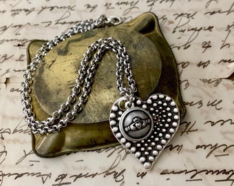 May include: A silver chain necklace with a heart-shaped pendant featuring a Celtic knot design. The pendant is textured with small beads.