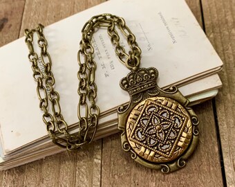 May include: A gold-toned necklace with a large pendant featuring a crown and intricate design. The pendant has a square center with a floral pattern and is surrounded by a decorative border.