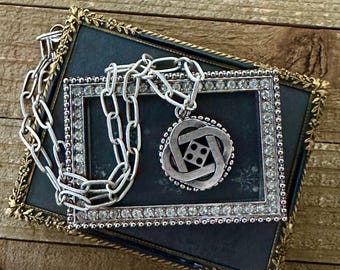 May include: A silver-toned necklace with a pendant featuring an interwoven design and a central square with four dots. The necklace rests on a dark blue framed picture with a silver border and clear stones. The frame has a gold trim.
