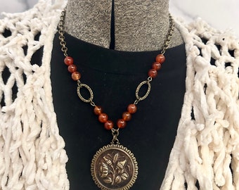 May include: A necklace with a large, round pendant featuring a floral design. The pendant is made of brass and has a textured surface. The necklace is made of brown beads and a chain.