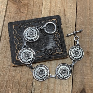 May include: A silver bracelet with a toggle clasp and five round, silver charms featuring a floral design.