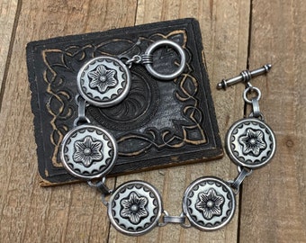 May include: A silver bracelet with a toggle clasp and five round, silver charms featuring a floral design.