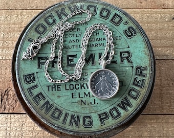 May include: A silver chain necklace with a round pendant featuring a stylized bee design. The necklace is resting on a vintage tin container with the text "Lockwood's Premier Blending Powder" printed on it.