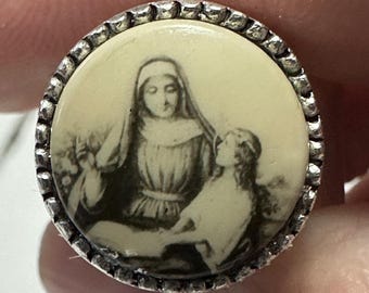 May include: A silver-toned pendant with a white cameo featuring a black and white image of a woman and a child. The woman is wearing a long robe and a head covering. The child is looking up at the woman.