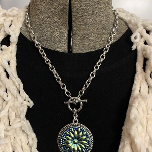 May include: A silver chain necklace with a toggle clasp and a round pendant featuring a blue and green flower design.