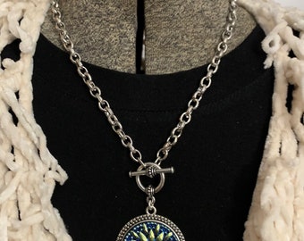 May include: A silver chain necklace with a toggle clasp and a round pendant featuring a blue and green flower design.