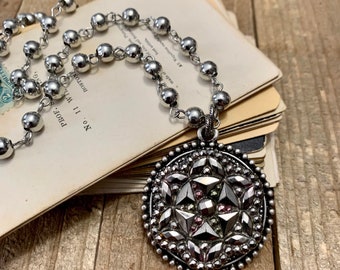May include: A silver necklace with a large, round pendant. The pendant is made of silver and has a geometric design with pink and green accents. The necklace is made of silver beads.