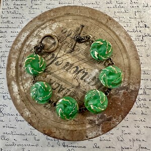 May include: A green glass bracelet with gold accents and a brass clasp. The bracelet is made of round beads with a leaf design. The bracelet is lying on a metal surface.