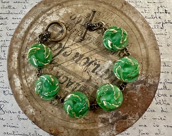 May include: A green glass bracelet with gold accents and a brass clasp. The bracelet is made of round beads with a leaf design. The bracelet is lying on a metal surface.