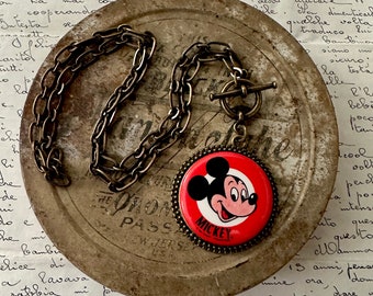 May include: A gold chain necklace with a round pendant featuring a cartoon image of Mickey Mouse. The pendant has a red background with a white outline and the word "MICKEY" printed in black at the bottom. The necklace is lying on a vintage tin container with handwritten text.