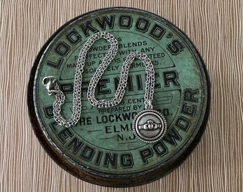 May include: A silver-toned necklace with a round pendant featuring a Claddagh design, resting on a vintage green tin. The tin has text that reads "LOCKWOOD'S BLENDING POWDER". The background is a brown and white striped pattern.