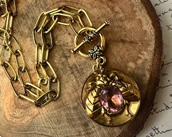 May include: A gold chain necklace with a toggle clasp and a pendant featuring a stylized bee with a large pink gemstone in the center.