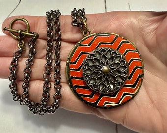 May include: A silver chain necklace with a large, round pendant. The pendant is made of metal and has a black and orange zig-zag pattern with a silver filigree design in the center.