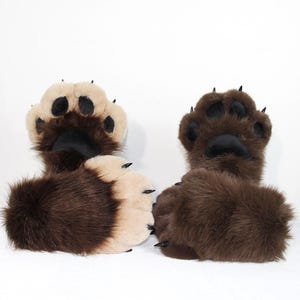 Puede incluir: Un par de pantuflas de pata de animal hechas de piel sintética marrón oscuro, con almohadillas de color beige claro y garras negras. Las pantuflas tienen una forma redondeada, una textura suave y costuras detalladas en los dedos.