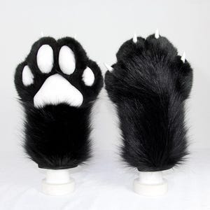 Op de afbeelding: Een paar zwarte imitatiebont handschoenen ontworpen als dierenpoten. De ene handschoen toont witte gevoerde zooltjes, terwijl de andere de achterkant laat zien met puntige witte klauwen die uit de vingertoppen steken. De textuur is lang.