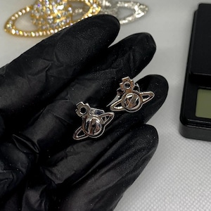 May include: A pair of silver-toned orb-shaped stud earrings featuring a cross emblem at the top and a ring encircling the center. In the background, two larger orb-shaped brooches in gold and silver tones are visible next to a digital scale.