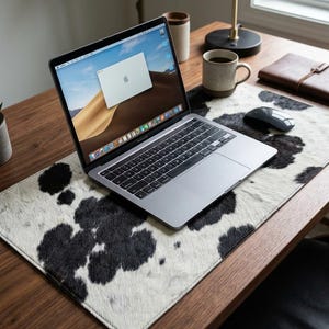 May include: A rectangular desk mat featuring a black and white cowhide pattern with a textured surface. The mat is positioned on a wooden desk surface and supports a silver laptop, a black computer mouse, and a ceramic coffee mug.