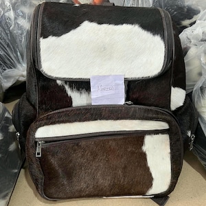 May include: A backpack made of cowhide with a brown and white spotted pattern. The bag features a large main compartment with a flap closure and a smaller front pocket with a zip. A small white paper tag with the handwritten name Marzia is attached.