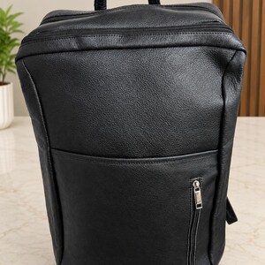 May include: A black leather backpack with a textured surface, featuring a top handle, a main zippered compartment, and a smaller front pocket with a vertical silver zipper closure. The design includes a clean, structured silhouette with visible stitching.