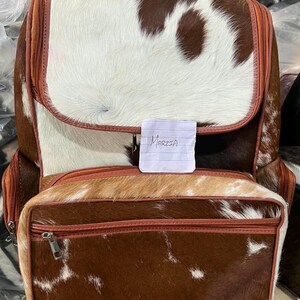 May include: A backpack featuring a brown and white cowhide pattern with a textured fur finish. The bag includes a large main compartment with a flap, a front zippered pocket, and brown leather trim. A small white paper note with the text Marzia is attached.