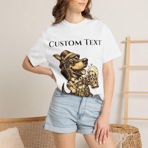 Könnte beinhalten: Ein weißes T-Shirt mit kurzen Ärmeln und einer braunen Grafik eines Dackels, der einen traditionellen Alpenhut und eine Lederhose trägt und einen Bierkrug hält. Der Text CUSTOM TEXT ist in Schwarz über der Hundeabbildung gedruckt.
