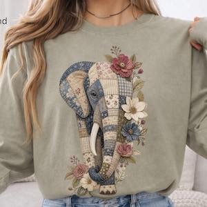 Puede incluir: Una sudadera de manga larga color arena con un gran gráfico de un elefante compuesto por varios patrones de patchwork en tonos azules, beige y marrones. El elefante está decorado con varias ilustraciones florales en rosa, blanco y azul.