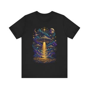 May include: A black t-shirt featuring a graphic print of multiple flying saucers hovering over a rural landscape at night. The design includes a bright yellow light beam, planets, stars, a crescent moon, and purple clouds in a celestial scene.