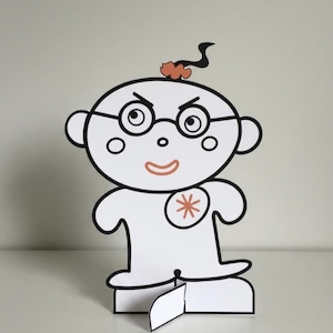 May include: A white paper cutout figure with black outlines, featuring a round head, glasses, and a small orange tuft of hair. The character has an orange mouth and an orange asterisk symbol on its chest, standing upright on a slotted base.