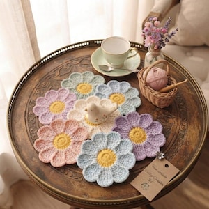 Op de afbeelding: Een set van zes gehaakte bloemonderzetters in pasteltinten roze, lavendel en blauw, elk met een geel hart. Een klein keramieken vogelbeeldje staat in het midden van de onderzetters, vergezeld door een label Handmade Daisy Coaster Set.