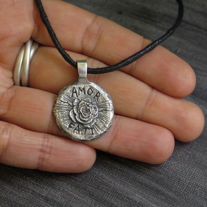 May include: Silver pendant necklace with a black cord. The pendant is round and features a rose with the words "AMOR FATI" inscribed around it.