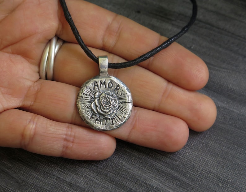 May include: Silver pendant necklace with a black cord. The pendant is round and features a rose with the words "AMOR FATI" inscribed around it.