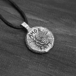 May include: A silver pendant necklace with a round charm featuring a rose and the words "AMOR FATI" in a stylized font.