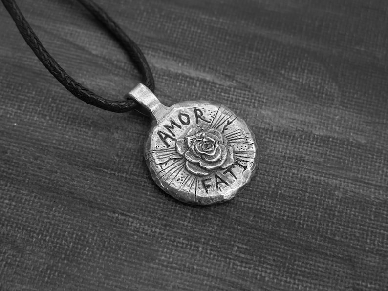 May include: A silver pendant necklace with a round charm featuring a rose and the words "AMOR FATI" in a stylized font.