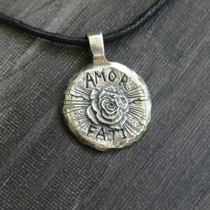 May include: Silver pendant necklace with a round charm featuring a rose and the words "AMOR FATI" in a stylized font.