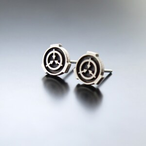 May include: A pair of silver earrings with a black and white symbol. The symbol is a circle with three arrows pointing inwards.
