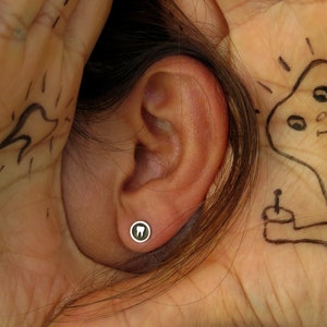 Puede incluir: Primer plano de la oreja de una persona con un pequeño pendiente de plata en forma de diente. El pendiente está en el lóbulo inferior de la oreja.