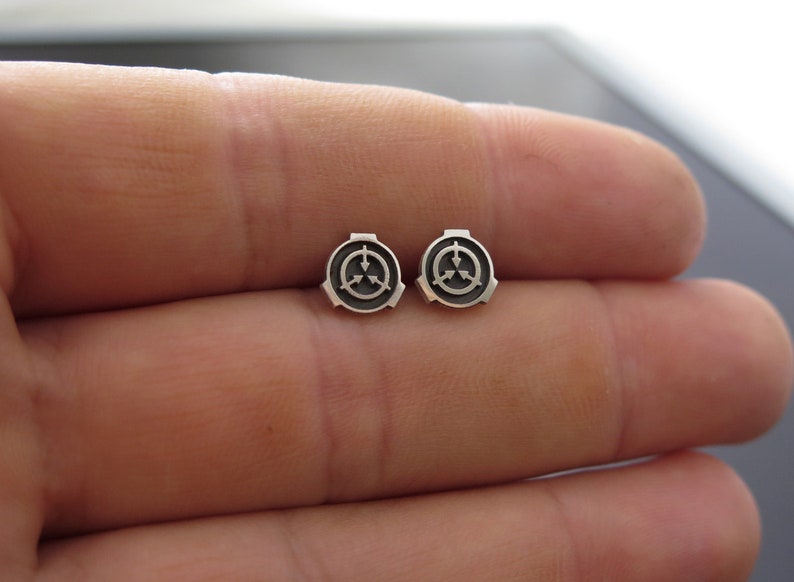 May include: A pair of silver earrings featuring a black and white symbol. The symbol is a circle with three arrows pointing inward.