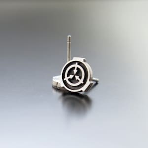 May include: A silver earring with a black circular design featuring three arrows pointing inwards. The earring is on a white surface.