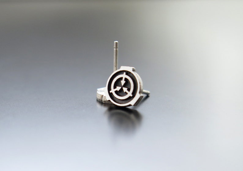 May include: A silver earring with a black circular design featuring three arrows pointing inwards. The earring is on a white surface.