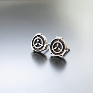 May include: A pair of silver stud earrings featuring a black and white symbol with three arrows pointing in different directions. The symbol is surrounded by a silver ring.